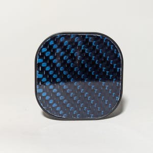 May include: A black square-shaped button with a blue and black carbon fiber pattern.