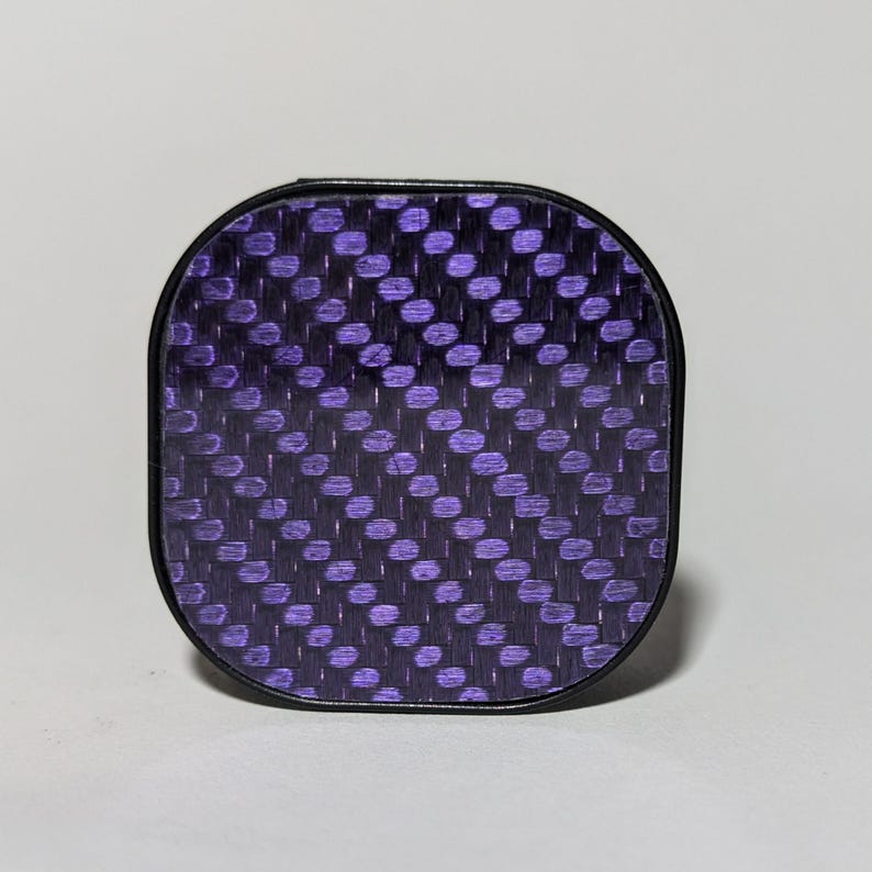 May include: A black square-shaped phone case with a purple carbon fiber pattern.