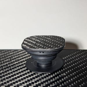 May include: A black phone grip with a square top featuring a carbon fiber pattern.