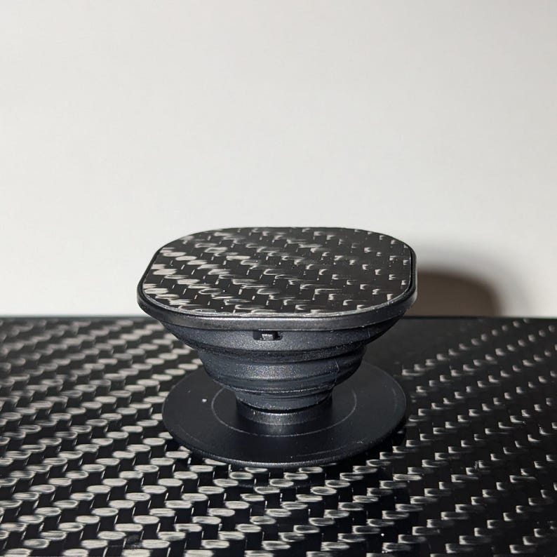May include: A black phone grip with a square top featuring a carbon fiber pattern.