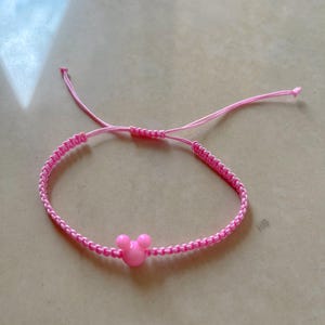 May include: Pink adjustable bracelet featuring a Mickey Mouse head charm. The bracelet is made of woven pink cord and has a sliding knot closure. The charm is a solid pink color.
