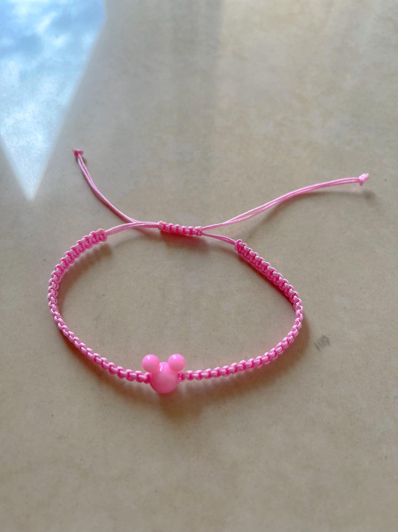 May include: Pink adjustable bracelet featuring a Mickey Mouse head charm. The bracelet is made of woven pink cord and has a sliding knot closure. The charm is a solid pink color.