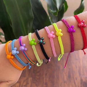 Könnte beinhalten: Eine Sammlung bunter Freundschaftsarmbänder mit Mickey-Mouse-förmigen Anhängern. Die Armbänder sind in verschiedenen Farben erhältlich, darunter Orange, Blau, Rosa, Grün, Schwarz, Gelb, Lila und Rot. Jedes Armband besteht aus geflochtenem Kordel.