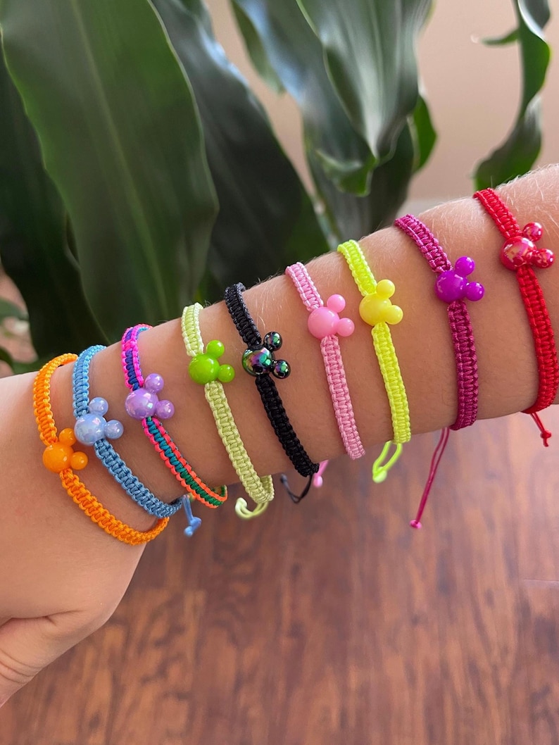 May include: A collection of colorful friendship bracelets with Mickey Mouse-shaped charms. The bracelets are in various colors, including orange, blue, pink, green, black, yellow, purple, and red. Each bracelet is made of woven cord.