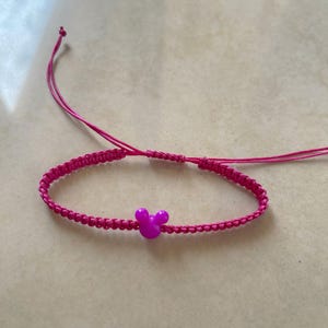 May include: A bright pink macrame bracelet with a small, purple Mickey Mouse head charm. The bracelet is adjustable with sliding knots and thin pink cords. The bracelet is on a light beige surface.