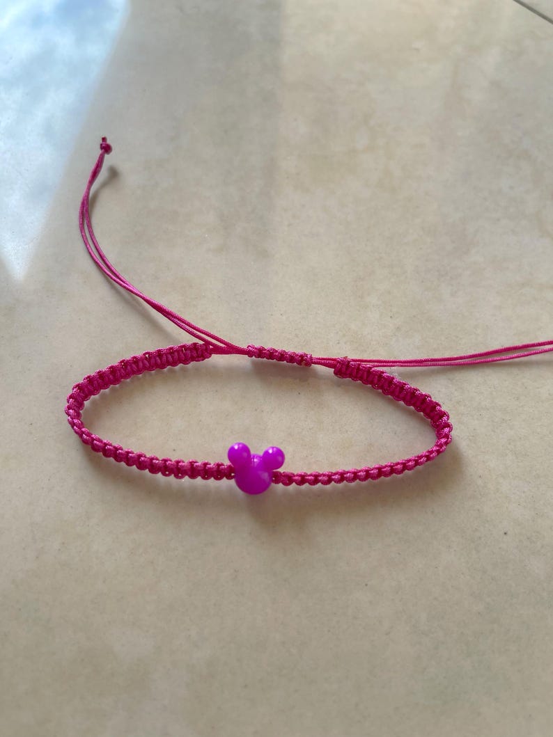 May include: A bright pink macrame bracelet with a small, purple Mickey Mouse head charm. The bracelet is adjustable with sliding knots and thin pink cords. The bracelet is on a light beige surface.