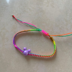 May include: A colorful friendship bracelet with a purple Mickey Mouse head charm. The bracelet is made of woven threads in a rainbow of colors, including pink, orange, green, and yellow. The adjustable closure features pink and orange threads.