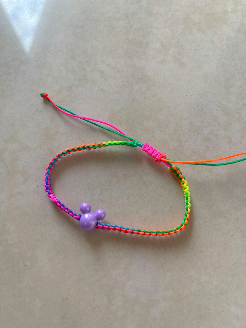 May include: A colorful friendship bracelet with a purple Mickey Mouse head charm. The bracelet is made of woven threads in a rainbow of colors, including pink, orange, green, and yellow. The adjustable closure features pink and orange threads.