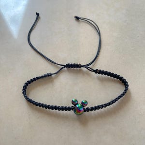 May include: Black braided bracelet featuring a Mickey Mouse head charm with iridescent coloring. The bracelet has an adjustable sliding knot closure. A fun accessory for Disney fans.
