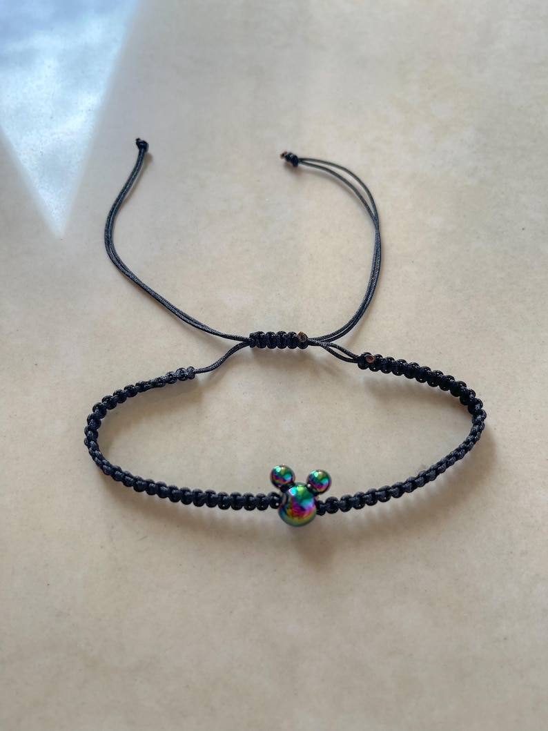 May include: Black braided bracelet featuring a Mickey Mouse head charm with iridescent coloring. The bracelet has an adjustable sliding knot closure. A fun accessory for Disney fans.
