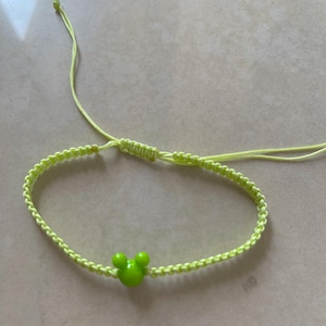 May include: A neon green macrame bracelet featuring a green Mickey Mouse head charm. The bracelet is adjustable with sliding knots and is made of a woven cord. The bracelet is a fun accessory.