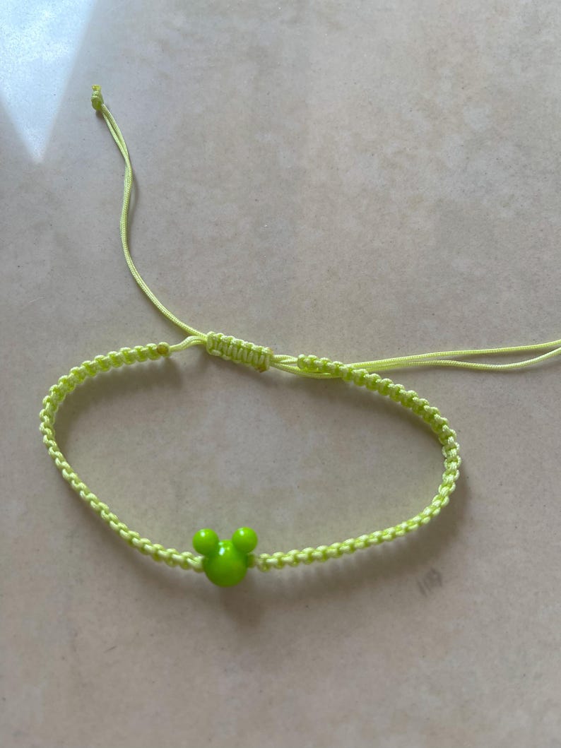 May include: A neon green macrame bracelet featuring a green Mickey Mouse head charm. The bracelet is adjustable with sliding knots and is made of a woven cord. The bracelet is a fun accessory.