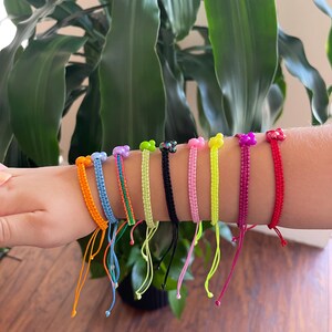 May include: A collection of colorful friendship bracelets. Each bracelet features a woven design in a different color, including orange, blue, green, black, pink, yellow, magenta, and red. The bracelets are adorned with small, decorative beads.