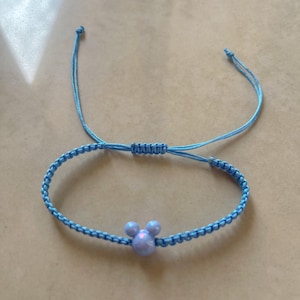 May include: A light blue adjustable bracelet featuring a Mickey Mouse-shaped charm. The bracelet is made of woven cord and has a sliding knot closure. The charm is a light purple color.
