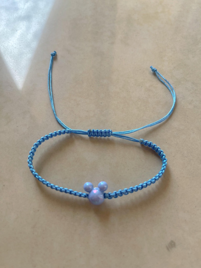 May include: A light blue adjustable bracelet featuring a Mickey Mouse-shaped charm. The bracelet is made of woven cord and has a sliding knot closure. The charm is a light purple color.