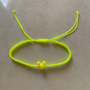 May include: Neon yellow adjustable bracelet featuring a yellow Mickey Mouse head charm. The bracelet is made of woven cord and has sliding knots for adjusting the size. A bright and cheerful accessory.