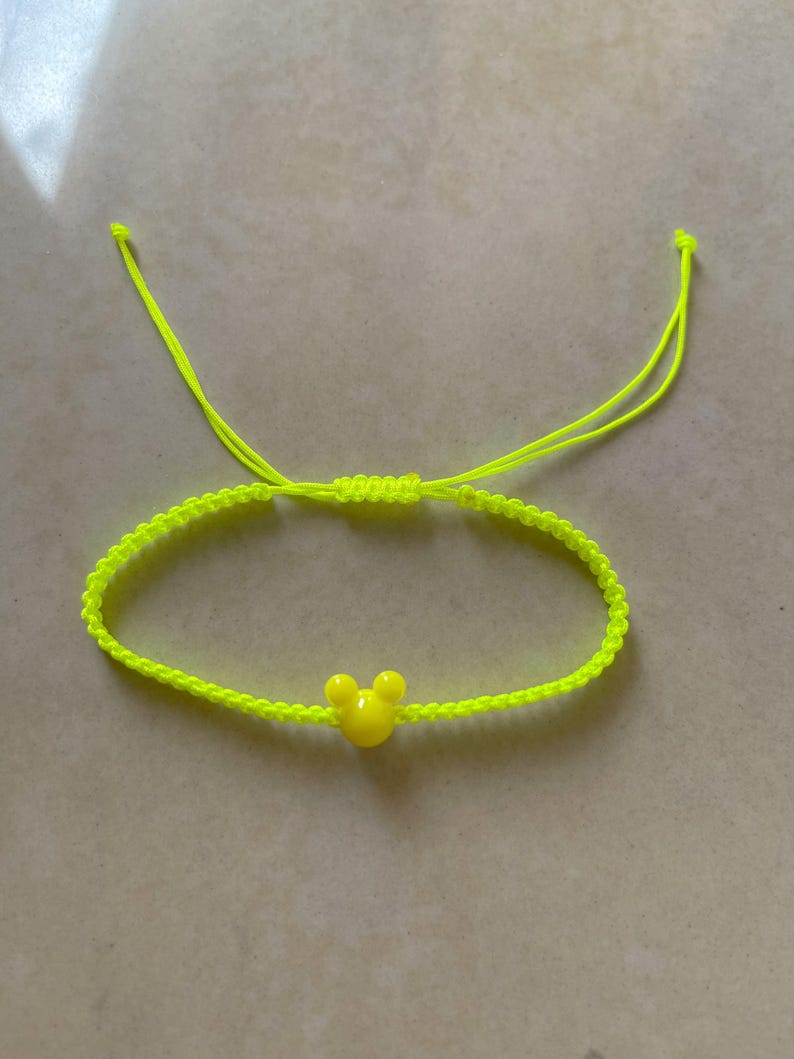 May include: Neon yellow adjustable bracelet featuring a yellow Mickey Mouse head charm. The bracelet is made of woven cord and has sliding knots for adjusting the size. A bright and cheerful accessory.