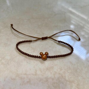 May include: Brown adjustable bracelet featuring a small, orange-brown Mickey Mouse head charm. The bracelet is made of a woven, dark brown cord with a sliding knot closure for an adjustable fit. The charm is centered on the bracelet.