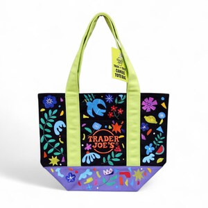 May include: Black canvas tote bag with a colorful floral and bird design. The bag has lime green handles and accents, with the Trader Joe's logo. A yellow tag reads "Trick or Treat Canvas Tote Bag."