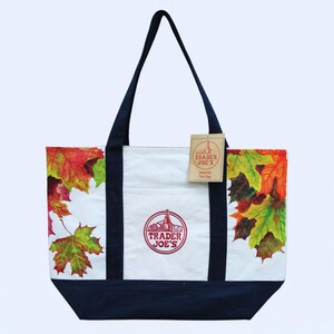 May include: A Trader Joe's tote bag with a white center panel and navy blue base and handles. The bag features a colorful autumn leaves design on the sides. The Trader Joe's logo is visible on the front, along with a hang tag.