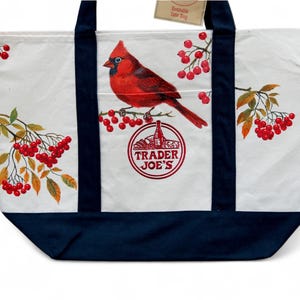 May include: White canvas tote bag with a navy blue base and straps. The bag features a vibrant red cardinal perched on a branch with red berries, and the Trader Joe's logo. The bag has a tag that says "Reusable Tote Bag".