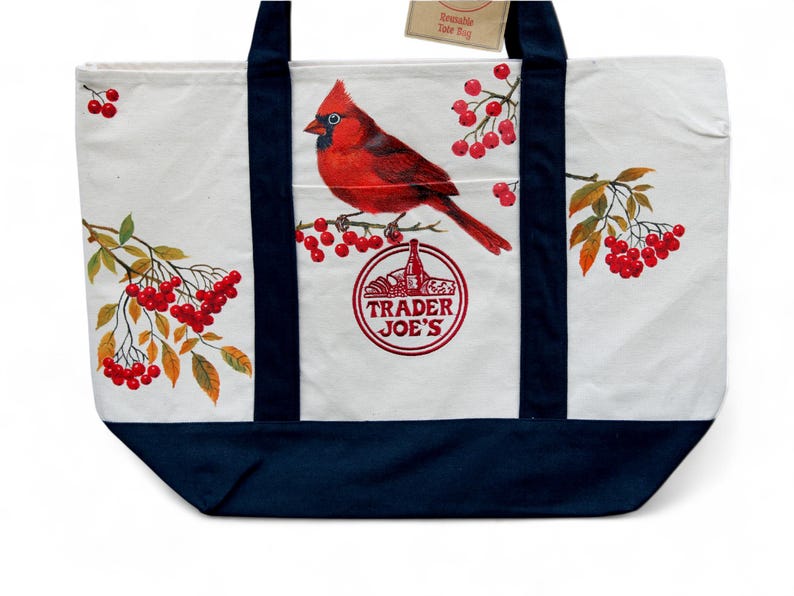 May include: White canvas tote bag with a navy blue base and straps. The bag features a vibrant red cardinal perched on a branch with red berries, and the Trader Joe's logo. The bag has a tag that says "Reusable Tote Bag".