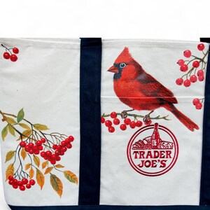 May include: White canvas tote bag with navy blue straps and trim. The bag features a vibrant red cardinal perched on a branch with red berries. The Trader Joe's logo is printed on the bag.