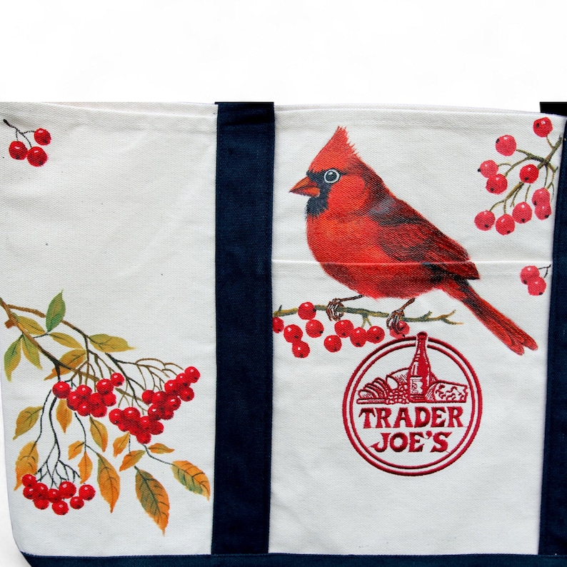 May include: White canvas tote bag with navy blue straps and trim. The bag features a vibrant red cardinal perched on a branch with red berries. The Trader Joe's logo is printed on the bag.