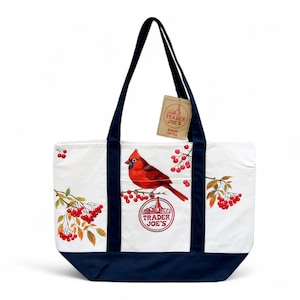May include: White tote bag with navy blue accents and handles. The bag features a red cardinal bird illustration with red berries and the text "Trader Joe's". A small tag with the Trader Joe's logo hangs from the handle.