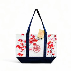 May include: White tote bag with a navy blue base and handles. The bag features a red maple leaf design and the Trader Joe's logo. A small tag is attached to the handle.