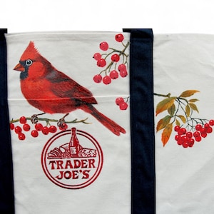 May include: White tote bag featuring a vibrant red cardinal perched on a branch with red berries. The bag has navy blue straps and a circular logo with the text "Trader Joe's" in red.