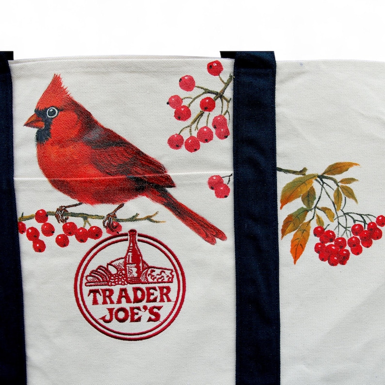 May include: White tote bag featuring a vibrant red cardinal perched on a branch with red berries. The bag has navy blue straps and a circular logo with the text "Trader Joe's" in red.