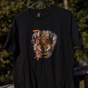 May include: Black t-shirt with a golden and orange dragon graphic. The dragon design features intricate details and is set against a smoky background. The shirt is hanging on a black hanger.