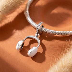 May include: A silver charm shaped like a pair of white headphones with a small silver bail featuring embedded clear stones, attached to a flexible silver snake chain bracelet. The charm has a polished metallic finish and white ear cups.