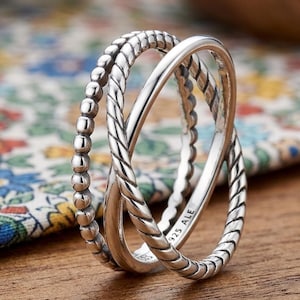 May include: A silver ring with an intertwined design. One band has a beaded texture, while the other two bands have a braided rope design. The ring is marked with "925 ALE". The ring is displayed on a wooden surface.