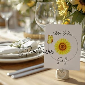 May include: A table setting with a white card featuring a yellow sunflower and butterfly design. The card reads "Table Yellow" and "S&J". The card is held by a heart-shaped holder. Other elements include plates, silverware, and floral arrangements.
