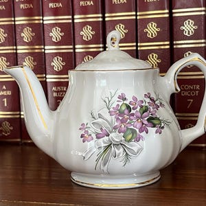 Könnte beinhalten: Eine weiße Teekanne mit einem Blumendesign aus lila Blüten und einer weißen Schleife. Die Teekanne hat einen goldenen Rand um den Rand und den Griff.