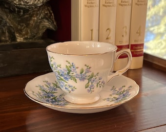 Könnte beinhalten: Eine weiße Teetasse und Untertasse mit einem floralen Muster aus blauen Vergissmeinnicht. Die Teetasse hat einen goldenen Rand und die Untertasse hat einen goldenen Streifen am Rand.