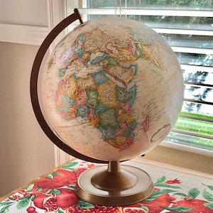 Może przedstawiać: Globus w stylu vintage z kremową powierzchnią, przedstawiający szczegółową mapę świata. Globus jest wsparty na stojaku w kolorze brązu i ciemnobrązowym pierścieniu południkowym. Stoi na stole z czerwonym kwiatowym wzorem.