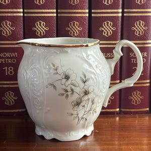 Peut inclure: Une crémière en porcelaine blanche avec un bord et une anse dorés. La crémière présente des motifs floraux et de volutes en relief, avec un motif floral peint dans des tons de gris et de brun. La crémière mesure environ 15 cm de haut.