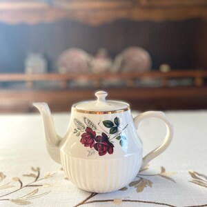 May include: Cream-colored teapot with a gold rim and lid. The teapot features a floral design with red roses and green leaves. The body has vertical ribbed detailing. The teapot is on a white tablecloth with a gold floral pattern.