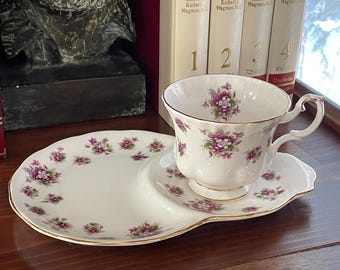 May include: A white teacup and saucer set with a floral pattern. The saucer has a small side plate for snacks. The teacup and saucer are decorated with a delicate floral pattern in shades of purple and green.
