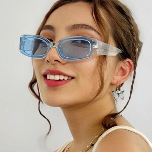 May include: Light blue rectangular sunglasses with a transparent frame. The image also shows a butterfly earring and layered necklaces. The person in the image is smiling.