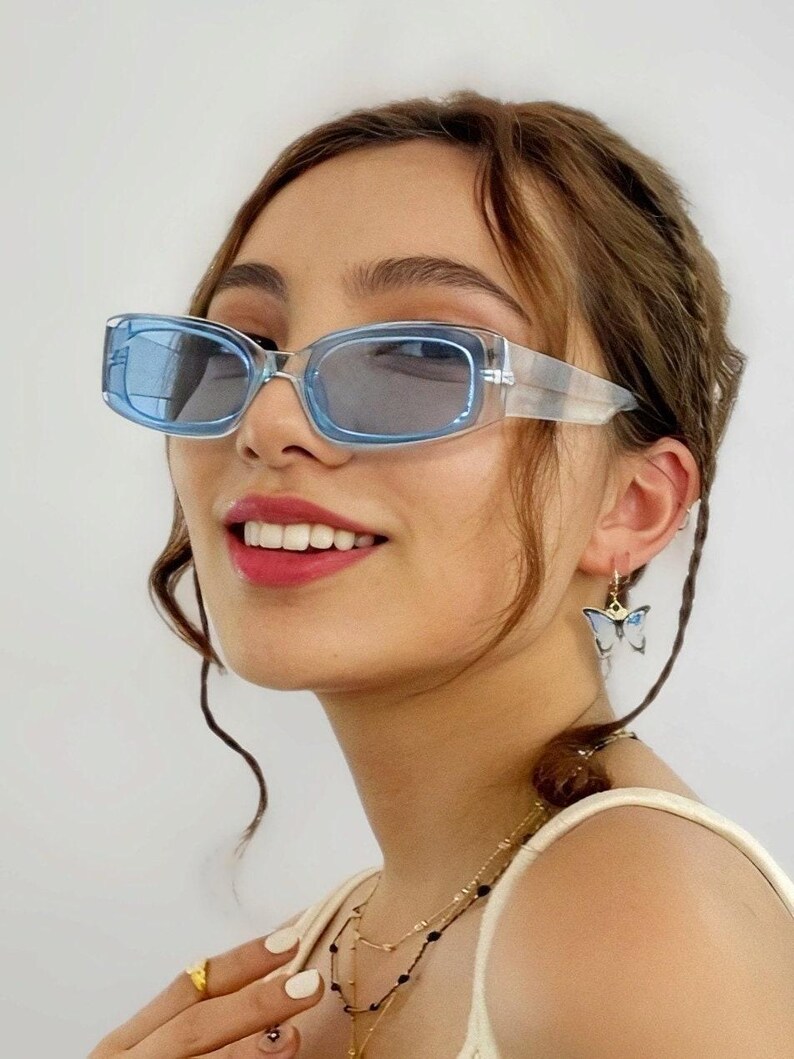 May include: Light blue rectangular sunglasses with a transparent frame. The image also shows a butterfly earring and layered necklaces. The person in the image is smiling.