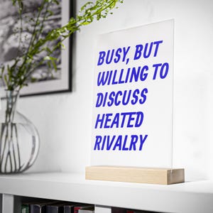 May include: A clear acrylic sign standing on a light-colored wooden base. The sign features bold blue text that reads: BUSY, BUT WILLING TO DISCUSS HEATED RIVALRY. The rectangular display is positioned upright on a flat surface.