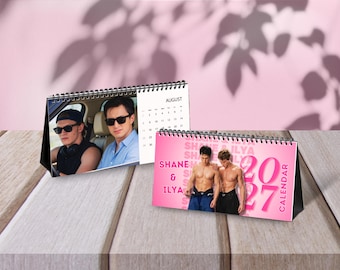 Peut inclure: Deux calendriers de bureau avec une reliure spirale noire. L'un montre une photo de deux personnes en voiture et le mois d'août. L'autre présente un fond rose avec le texte Shane & Ilya 2027 Calendar et une photo de deux personnes torse nu.
