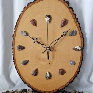 May include: A wooden clock with a round, natural wood face and black hands. The clock face is decorated with 12 arrowheads made of different types of stone.