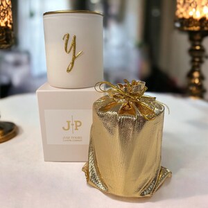 May include: A white candle with a gold rim and a gold letter "y" on the front, on a white box with "JP" printed on it. A gold gift bag is next to the candle. The candle and gift bag are on a white surface.