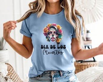 May include: A light blue t-shirt featuring a graphic of a sugar skull face adorned with colorful roses. Below the illustration, the text reads "DIA DE LOS Muertos" in a combination of bold, rounded block letters and a cursive script font.