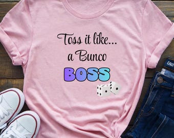 Könnte beinhalten: Hellrosa T-Shirt mit dem Text "Toss it like... a Bunco BOSS" in schwarzer und blauer Schrift. Zwei weiße Würfel mit schwarzen Punkten sind darunter abgebildet. Das T-Shirt ist unten geknotet und wird mit blauen Jeans und weißen Turnschuhen kombiniert.